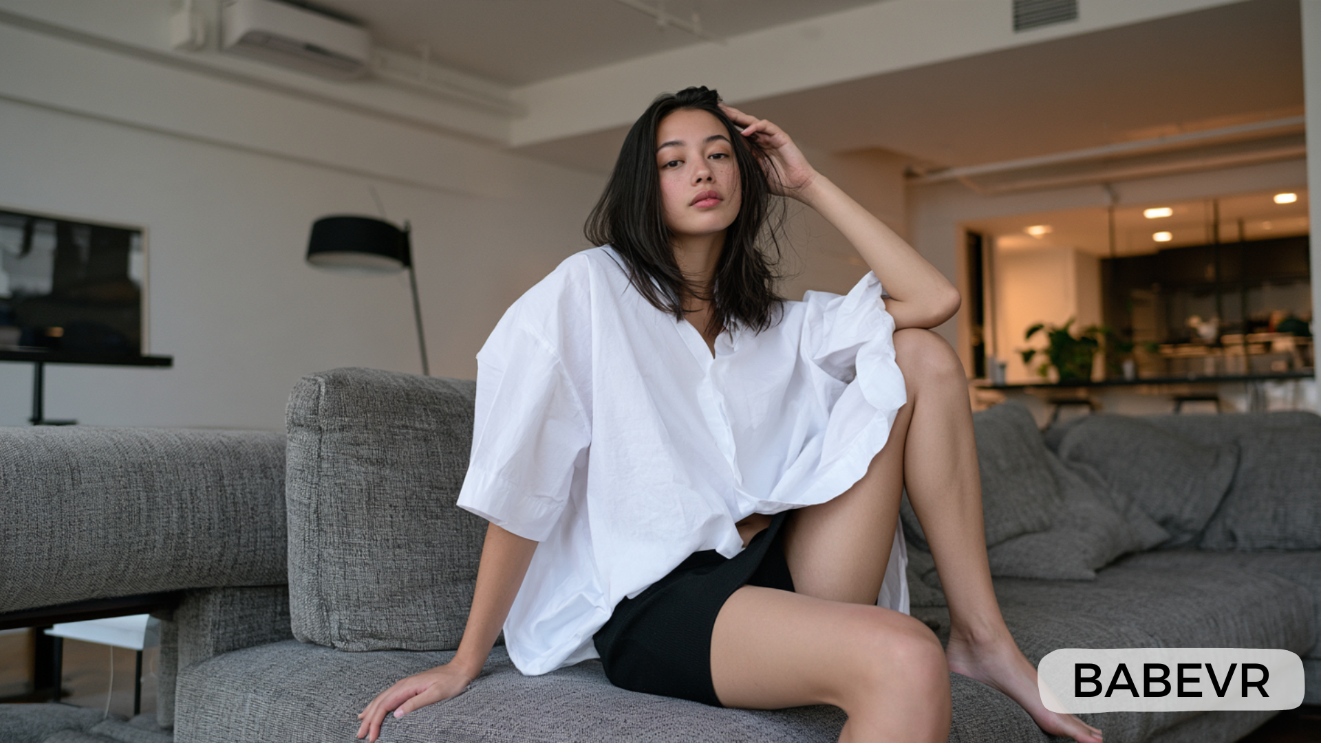 Femme adulte aux cheveux noirs mi-longs, vêtue d’une chemise blanche ample et d’un short noir, assise sur un canapé gris dans un intérieur moderne, avec une jambe relevée et le regard tourné vers la caméra
